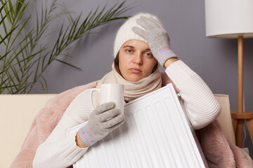 Image of sick woman in coat and hat sitting in cold living room with radiator, struggling from...