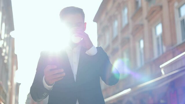 Handsome happy male professional typing on mobile phone while walking outdoor in good mood and drinking coffee. Caucasian joyful man texting on smartphone on street with coffee-to-go. Job concept