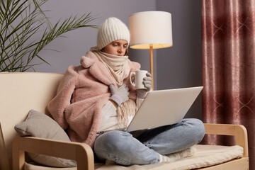 Profile portrait of woman freelancer in cold room sitting on the sofa wearing warm hat, mittens and cap, works on notebook and trying to get warm while working online on pc computer. © sementsova321