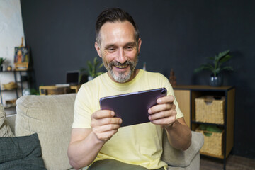 Playing online games middle aged grey haired man using digital tablet sitting on the sofa at home wearing casual. Mature freelancer man hold digital tablet work from home. Business online