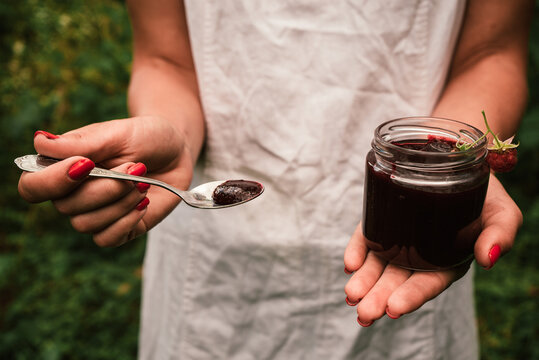 a woman in a white dress holds a jar and a spoon with berry or fruit jam in her hands 1
