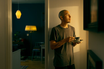 Fototapeta na wymiar A caucasian man with a shaved head standing in a room next to a kitchen in a warm light holding a vintage coffee cup.