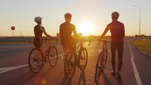 Two men and one woman in sport clothes and helmets chatting and smiling while walking with bikes outdoors. Young people enjoying summer sunset during outdoors workout.