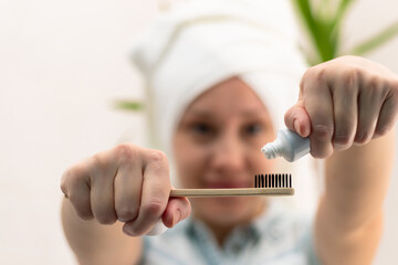 A young pretty woman with a towel on her head in a bright bathroom spreads toothpaste on a toothbrush against the background of a green plant. Selective focus. Close-up
