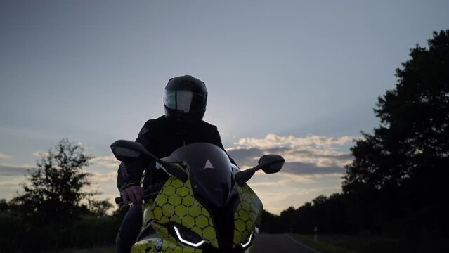 Biker man on BMW sport motorcycle, young man rider on trendy motorbike stopped on asphalt road in countryside to rest during the trip at sunset - orbiting gimbal shot