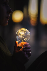 young worried female human holding bright shining lightbulb in her hands feeling unsave while thinking negative about a future blackout with no electricity or power outage with warm background