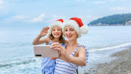 A family on the beach by the sea in Santa Claus Christmas hats communicates with relatives via phone.