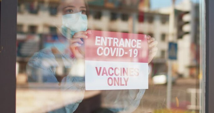 Business owner female wearing face mask attaching sign on front door glass, pandemic time. Woman attaching paper with text NEntrance Covid-19 vaccines only. Pandemic of coronavirus. Covid-19