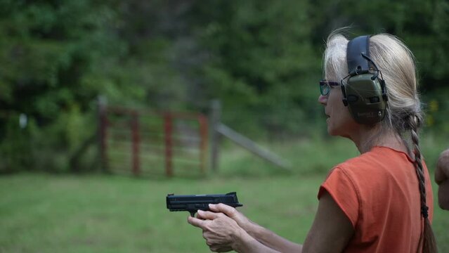 Mature, elderly woman fires a handgun at a shooting range target outside on a summer day.