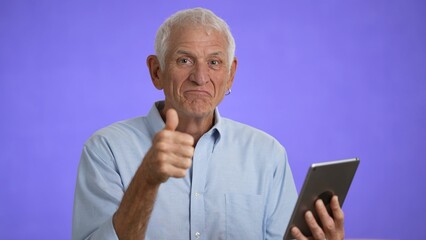 Happy elderly old man thumbs up with digital tablet computer, senior adult man enjoying using computer pad apps browsing internet shopping isolated on solid purple background.