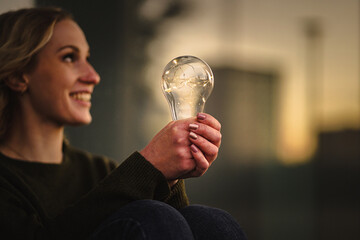 young smiling woman holding bright shining lightbulb in her hands while thinking optimistic about green alternative energy solution with solar light on a warm emotional background