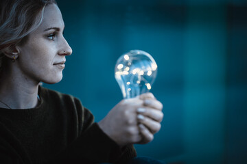 young worried woman holding bright shining lightbulb in her hands feeling unsave while in fear thinking about a future blackout with no electricity or power outage with blue cold background