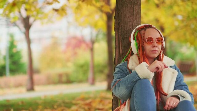 Lady with dreadlocks listening to music in park. Young woman with colorful dreadlocks in wireless headphones listening to music while leaning on tree trunk in autumn park