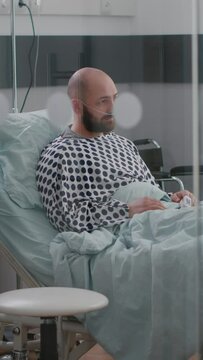 Vertical video: Stressed sick man sitting in bed waiting for respiratory treatment recovering after sickness surgery in hospital ward. Hospitalized patient looking into camera wearing nasal oxygen