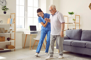 Young nurse helping elderly man walk in the room, holding his hand, supporting him. Treatment and rehabilitation after injury or stroke, life in assisted living facility, senior care concept - Powered by Adobe