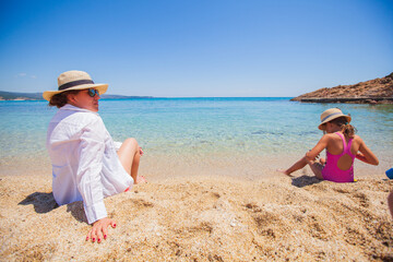Mother and little daughter relax on sand beach during summer holidays.
