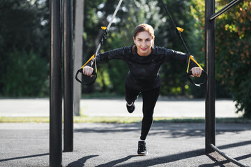 Women and sport. Smiling girl in sportswear with sports equipment TRX exercising outdoors at the...