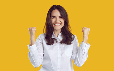 Happy satisfied woman clenches her fists feeling euphoric and rejoicing in her success. Joyful excited brunette Caucasian woman in white shirt rejoices on yellow background. Concept of luck.