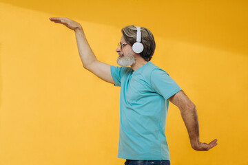 Optimistic gray-haired old man listens to music, dances, wears headphones, glasses, isolated on yellow background.