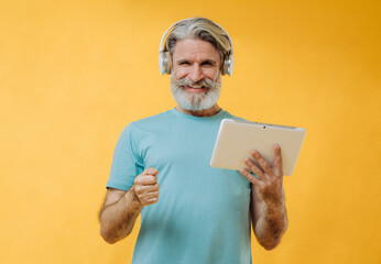 Photo of an expressive gray-haired senior man in headphones with a tablet in his hands, isolated on a yellow background.