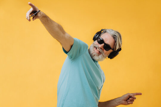 Photo Of An Expressive Gray-haired Senior Man In Headphones With A Phone In His Hands, Isolated On A Yellow Background.