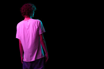 Back view of young hairy man, student in white tee isolated on dark background in neon light. Concept of emotions, facial expression, youth