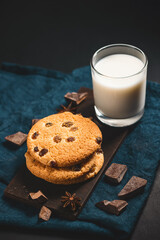 Crispy oatmeal cookies with raisins, glass with milk and chocolate chunks on dark background