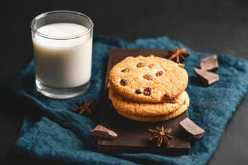 Crispy oatmeal cookies with raisins and glass with milk on wooden board, dark background