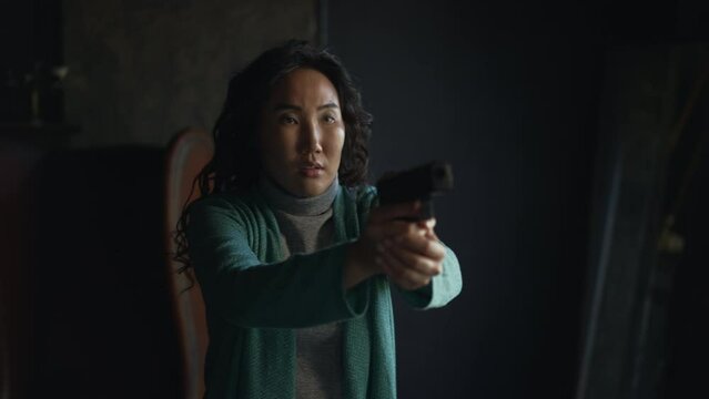 4k Young Asian woman is holding gun and standing in antique room spbd. Close view of beautiful curly female holds weapon in her hand and shows emotion excitement, stands against background of black