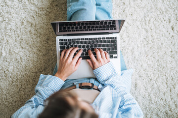 Fototapeta premium Crop photo of young woman in jeans working on laptop sitting on carpet at home, view from top