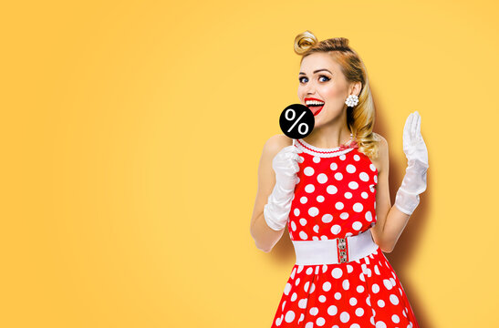 Very tasty discounts, rebates, deals concept image. Cheerful beautiful woman licking signboard with % sign, dressed in pinup red dress, isolated on orange yellow background. Black Friday sales ad.