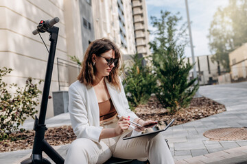 Young businesswoman in white suit sitting on electric scooter and working on digital tablet in city with modern architectury