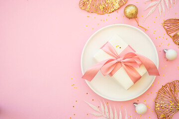 Christmas table setting with white plate and christmas golden decorations on pink background.