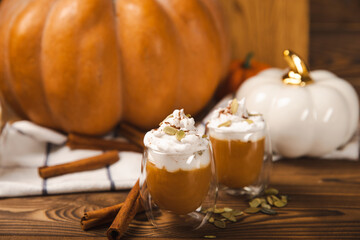 Pumpkin spice latte in glass mug with cinnamon, nutmeg, pumpkin seeds and whipped cream on brown texture wood. A glass of autumn coffee with pumpkin. A warming drink. Autumn concept.