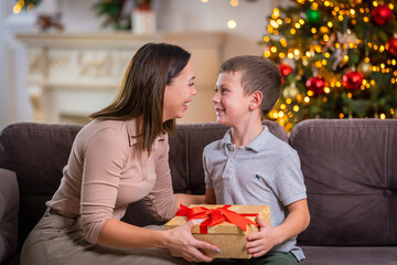 happy loving mother and son hugging at home, sitting on sofa against background Christmas tree. child and woman laughing happily, holding gift box in their hands. Give each other gifts at Christmas