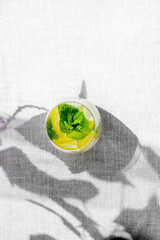 Top view of homemade lemonade with lemon, mint and ice cubes in glass on a grey background with hard leaves shadows.