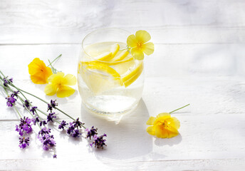 Homemade lemonade with lemon and ice cubes in a glass on the white wood table with yellow flowers and lavener
