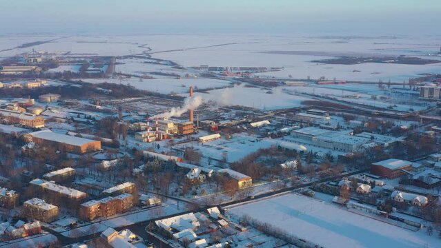 Above view, dolly move sideways, on industrial complex, smoke form chimneys of district heating plant, pollution, roofs of buildings, houses white of snow, frosty settlement, cityscape in winter.  