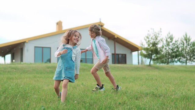Happy Caucasian small kids boy and girl playing outdoors running on grace in nature. Little children play outside the house in green field chasing each other. Happy careless childhood concept