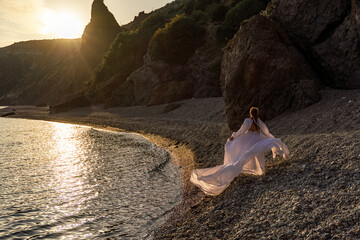 A mysterious female silhouette with long braids stands on the sea beach with mountain views, Sunset rays shine on a woman. Throws up a long white dress, a divine sunset.