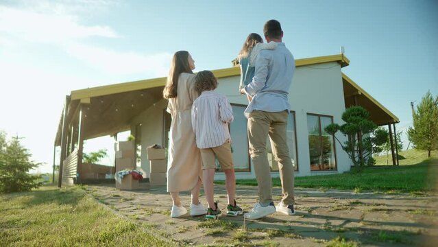 Back view of Caucasian joyful young family with son and daughter standing near new home outdoors looking at own house. Parents with children moving in new house. Settle down, boxes on background