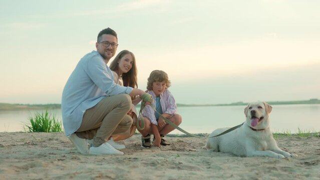 Positive young parents with child boy and cute dog sitting on a beach outdoors near lake smiling at camera. Family with son and pet near water together in good mood. family time. Weekend recreation