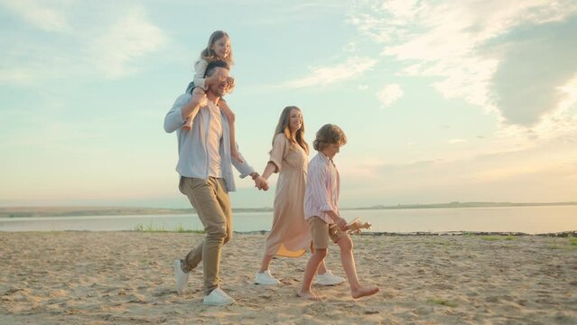 Happy married couple walking outdoor in nature with little children and smiling. Family with daughter and son walking on a beach near beautiful lake. Recreation. Summertime. Childhood concept