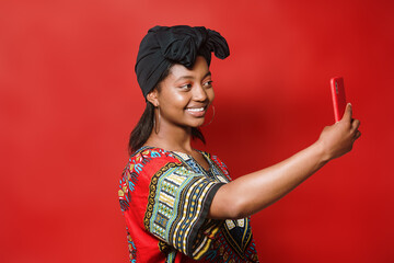 Beautiful young African American girl with a turban and typical dress taking a selfie with her phone on a red background