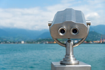 Metal gray stationary binoculars on the observation deck overlooking the sea, mountains and the city on a sunny day, copy space. Bizarre imaginary metal man