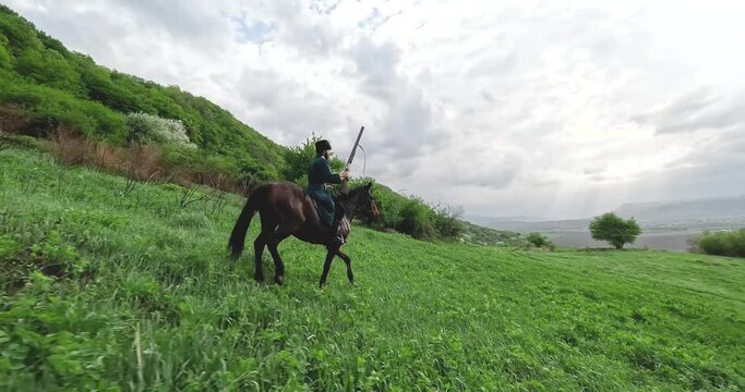 FPV sports drone shot traditional culture Caucasian horseman with gun riding horse at summer mountain valley landscape. Aerial view male equestrian highlander at picturesque dramatic sky rocky scenery