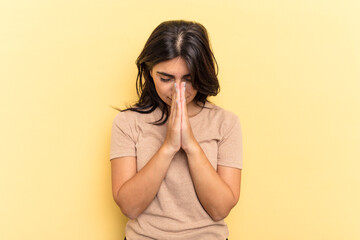 Young Indian woman isolated on yellow background praying, showing devotion, religious person looking for divine inspiration.