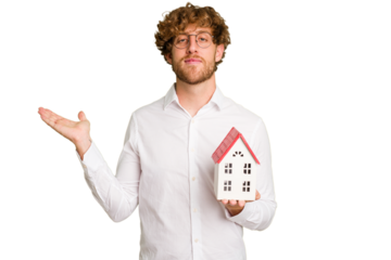 Business caucasian man holding toy house isolated on white background showing a copy space on a palm and holding another hand on waist.