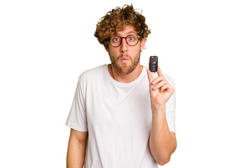 Young caucasian man holding car keys isolated on white background shrugs shoulders and open eyes confused.