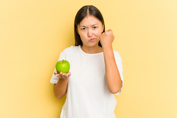 Young Asian woman eating an apple isolated on yellow background showing fist to camera, aggressive facial expression.